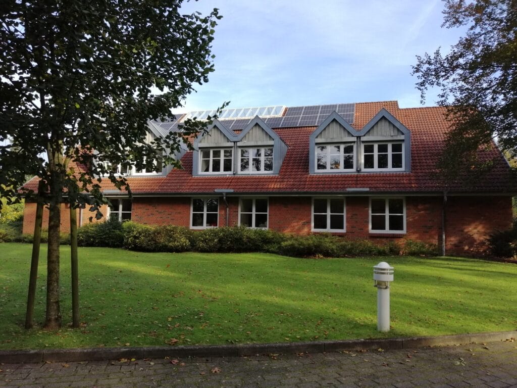 Das Verwaltungs-Gebäude der Stiftung Uhlebüll. Es ist ein rotes Backstein-Gebäude mit orange-roten Ziegeln und mehreren Gauben im Obergeschoss. Vor dem Haus ist eine Rasenfläche zu sehen.