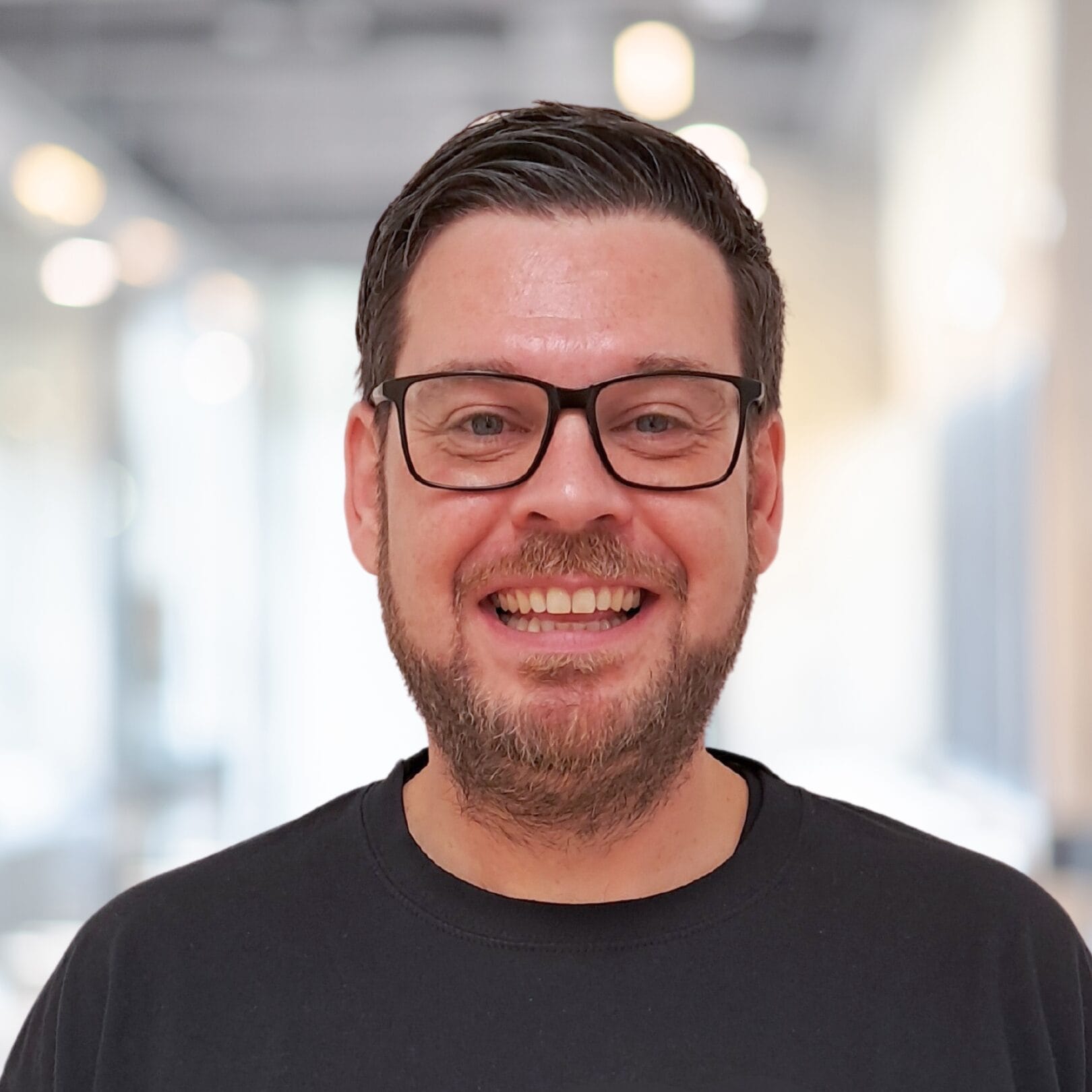 Ein Mann mittleren Alters lächelt in die Kamera. Er hat dunkles kurzes Haar, eine dunkle Brille und ein schwarzes Oberteil.